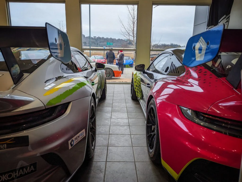 Two Porsche Race Cars with prominent rear spoilers, one silver and one red, inside a building. People can be seen outside through the
