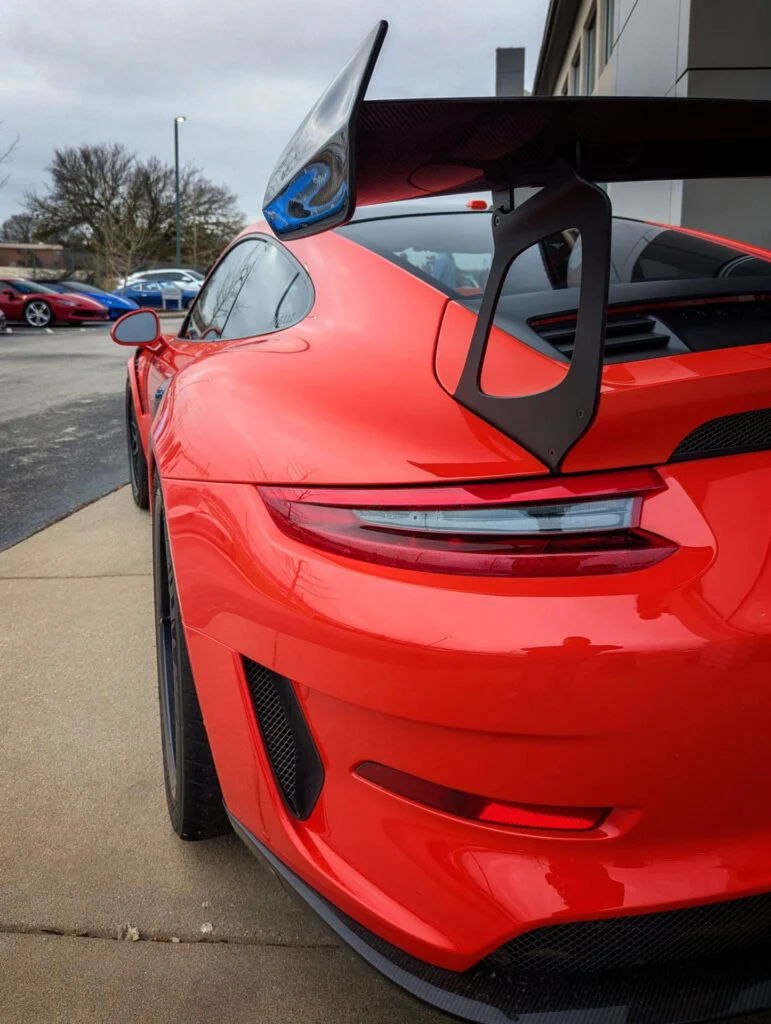 Porsche 911 GT3 RS with a large rear wing parked on a concrete surface, with other vehicles visible in the background.