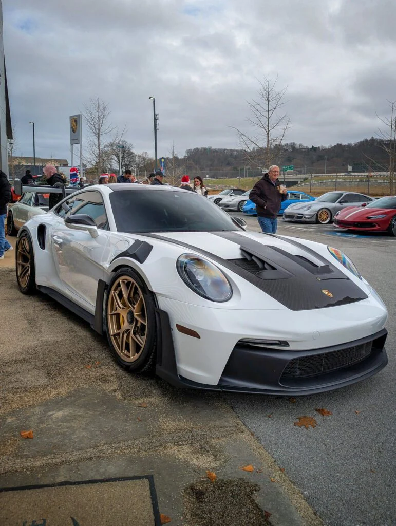 White Porsche 911 GT3 with gold wheels parked outside a dealership during an event with attendees in the background.