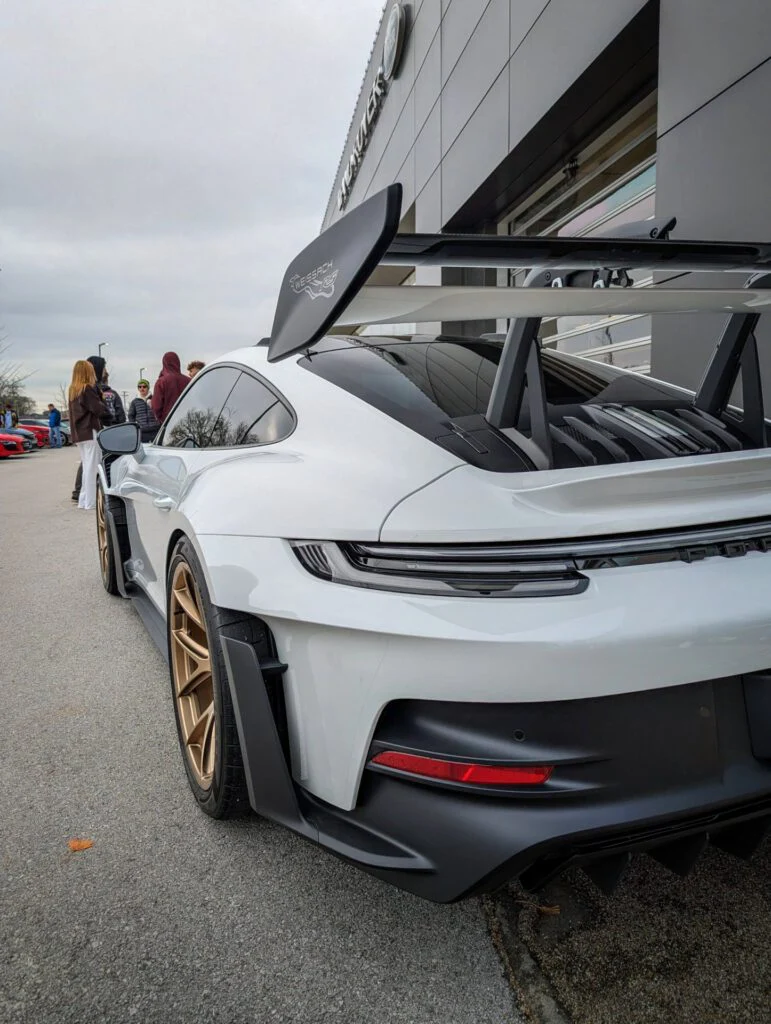 White Porsche 911 GT3 with a large rear wing parked near a building with attendees in the background.