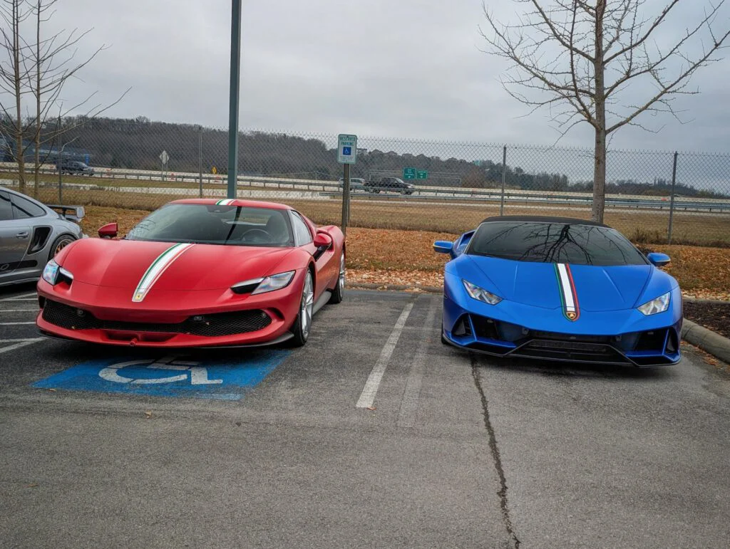 Red Ferrari and blue Lamborghini parked side by side in a lot, trees and cloudy sky in the background.