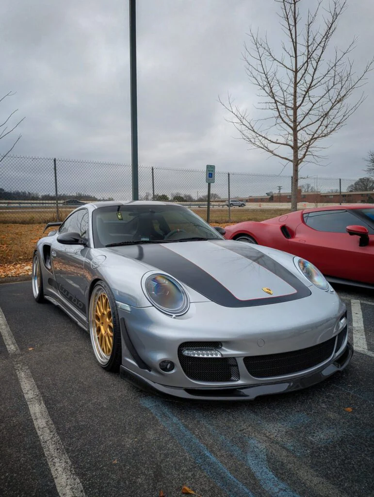 Silver Porsche 911 Turbo with aftermarket modifications parked in a lot next to a red sports car.