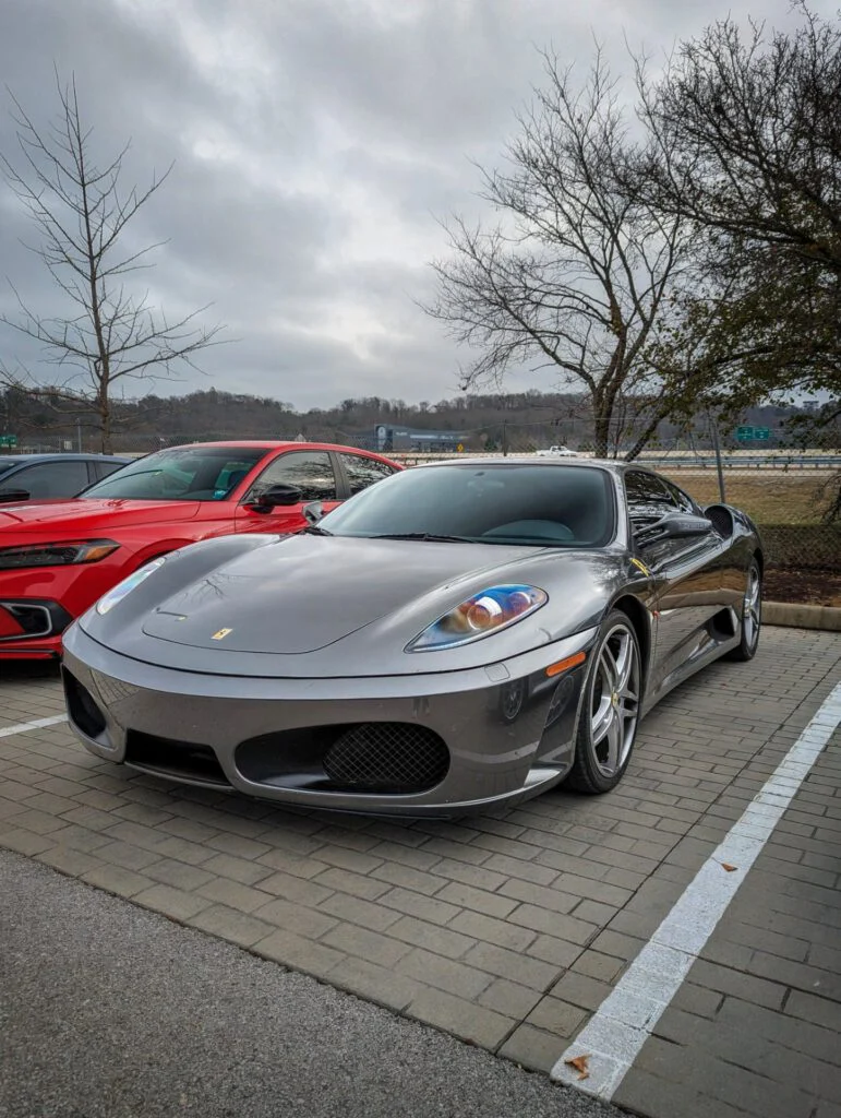 Gray Ferrari F430 parked next to a red car in a parking area with trees and a cloudy sky.