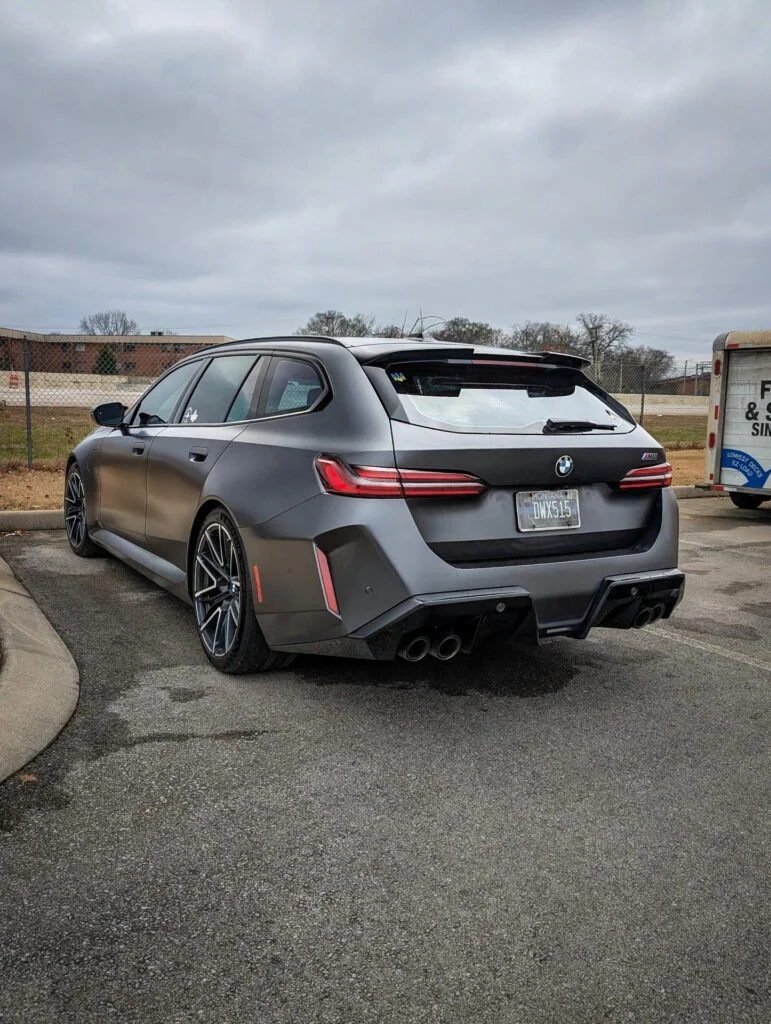 A gray BMW M5 Touring with distinctive rear design and dual exhaust parked in a lot under overcast skies.