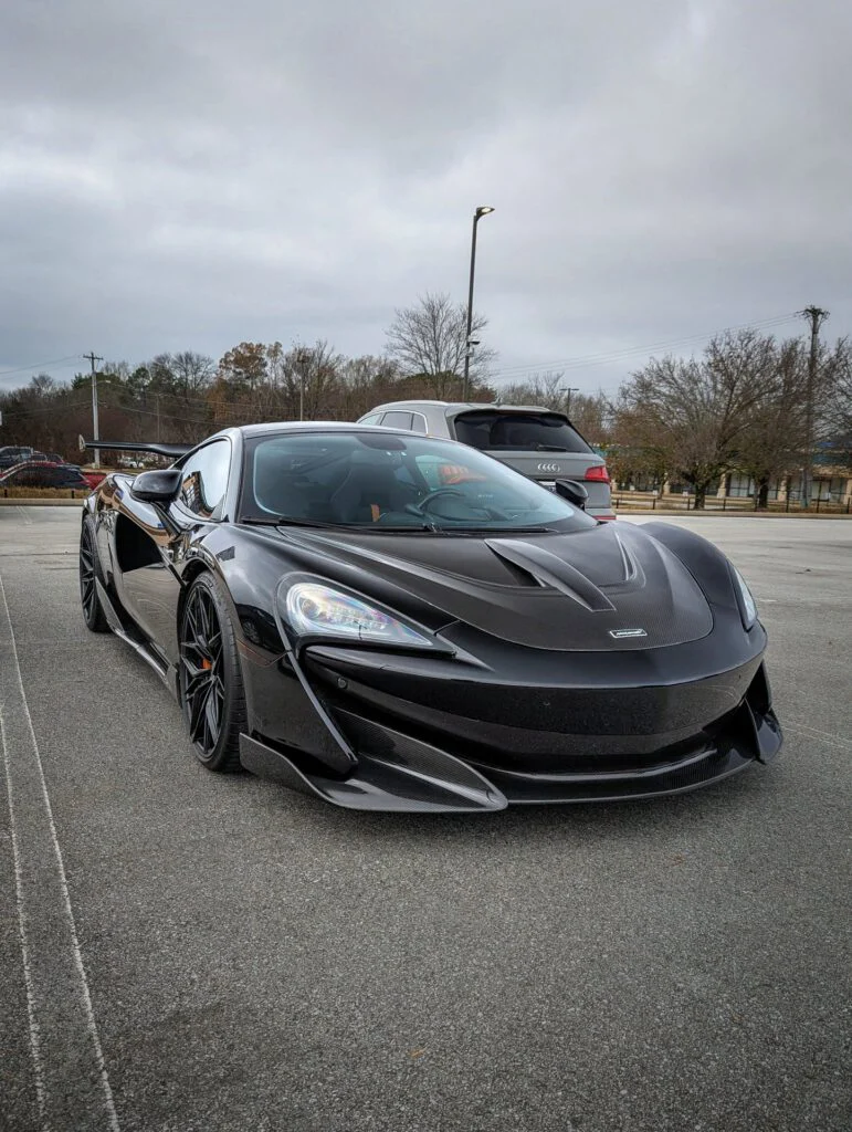A black McLaren 720S parked in a lot, with trees and a cloudy sky in the background.
