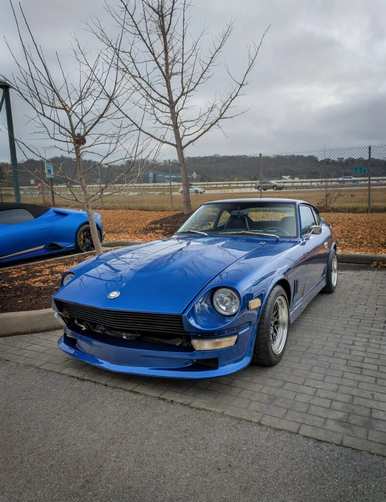 A blue Datsun Z parked on a paved area with trees and a highway in the background.