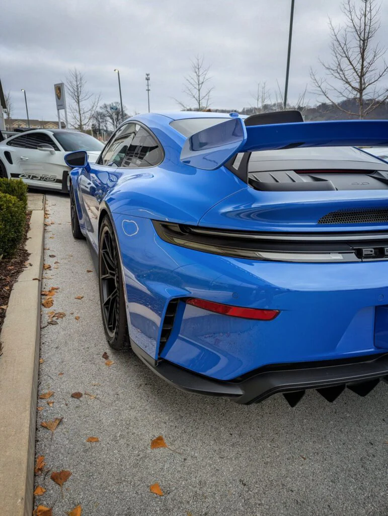 Blue Porsche 911 GT3 with a large wing next to a white car in a parking lot.