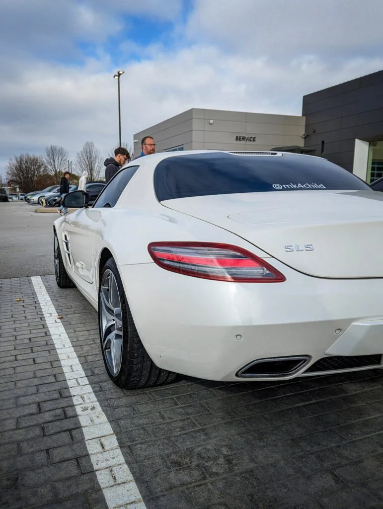 White Mercedes-Benz SLS AMG parked near a service area with individuals conversing in the background.