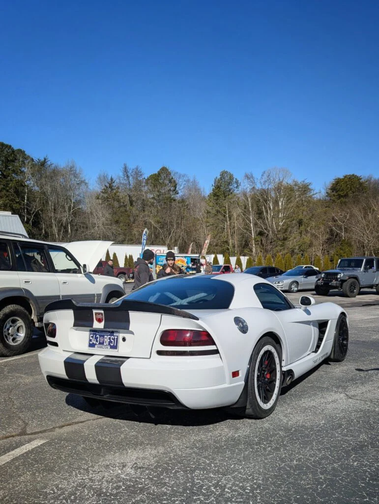 A white Dodge Viper with black stripes parked in a lot with people nearby.
