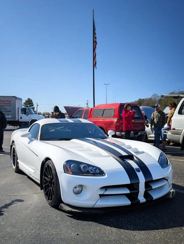 White Dodge Viper with black stripes parked among attendees at a car event, American flag in background.