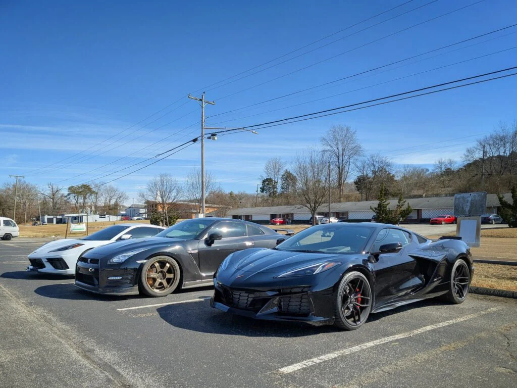 Three sports cars parked in a lot with a clear blue sky; Nissan GT-R, white Corvette, and black Corvette.