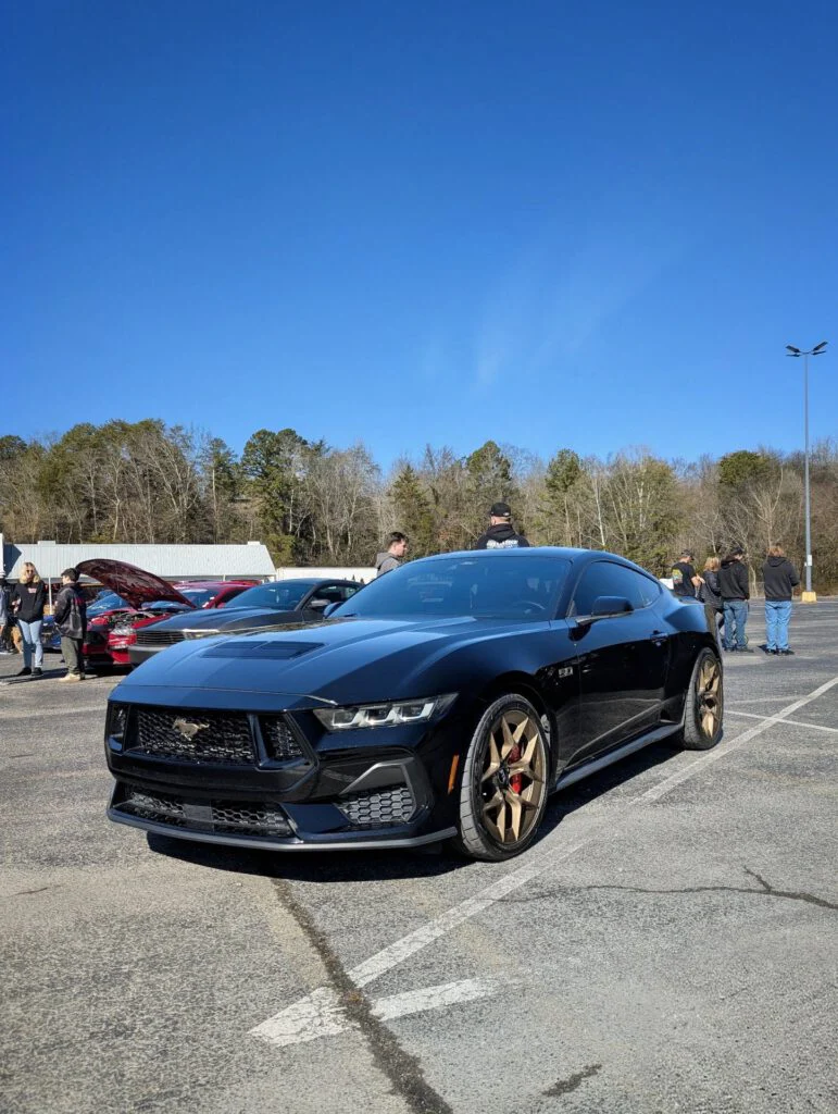 A black Ford Mustang parked in a lot with attendees in the background and clear blue skies.