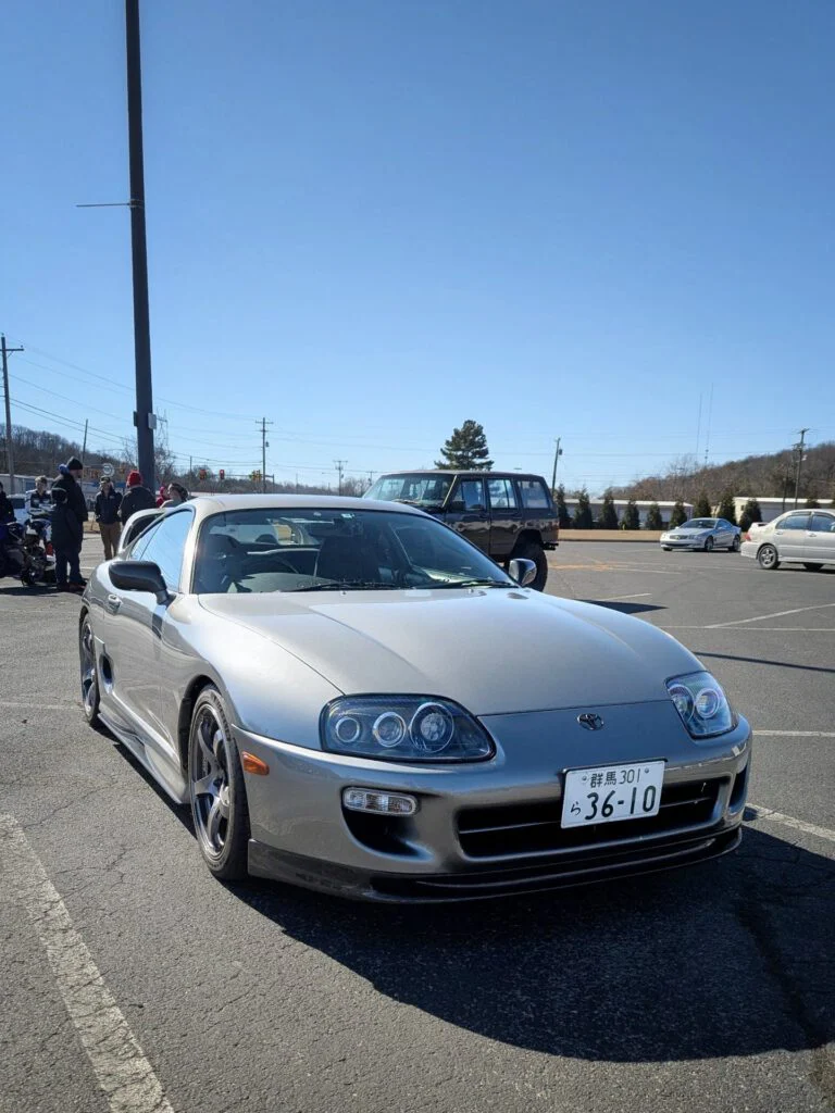 Silver sports car parked in an outdoor lot with people in the background.