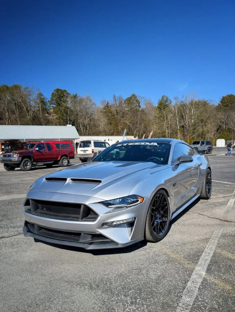 Silver Ford Mustang parked at an outdoor car event with trees in the background.