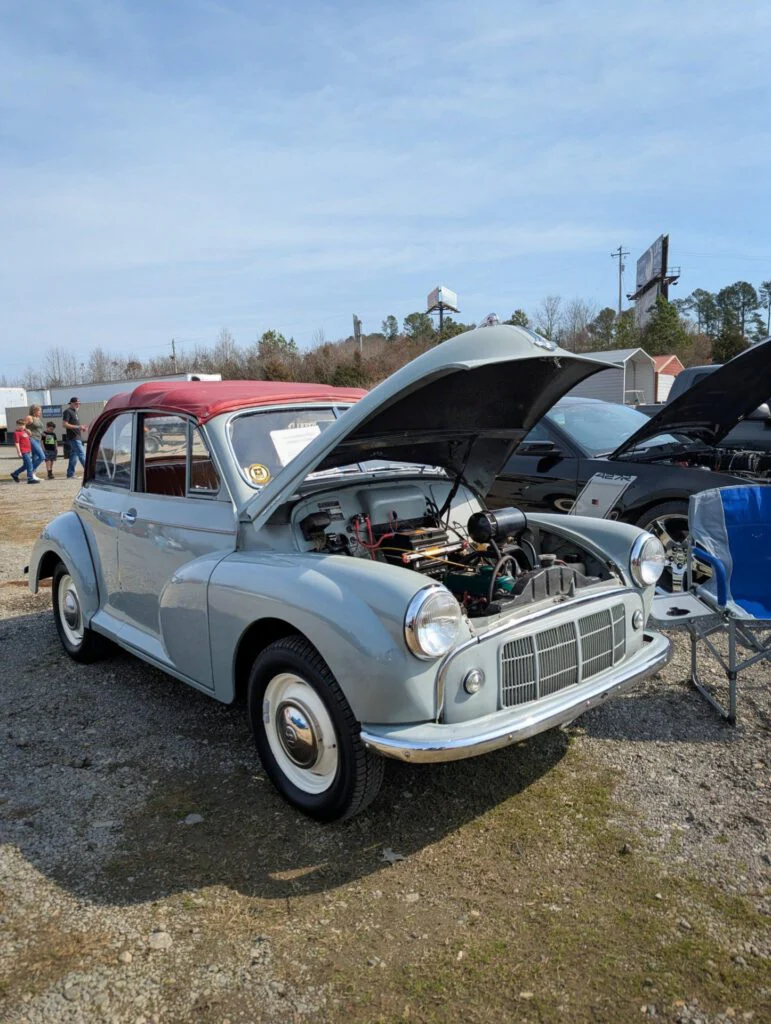 A light blue car with a red roof has its hood open, showcasing the engine. Attendees walk in the background.