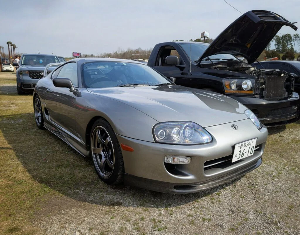 Silver Toyota Supra parked on grass with a black truck and SUV in the background.