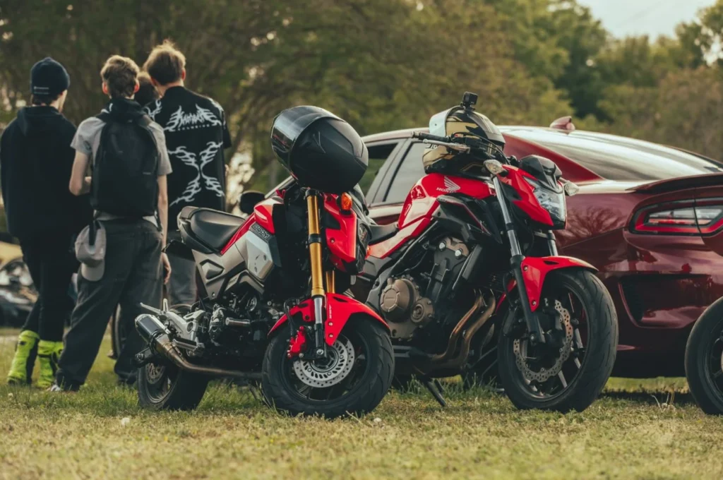 Red Honda motorcycle and red car at outdoor gathering with attendees nearby.