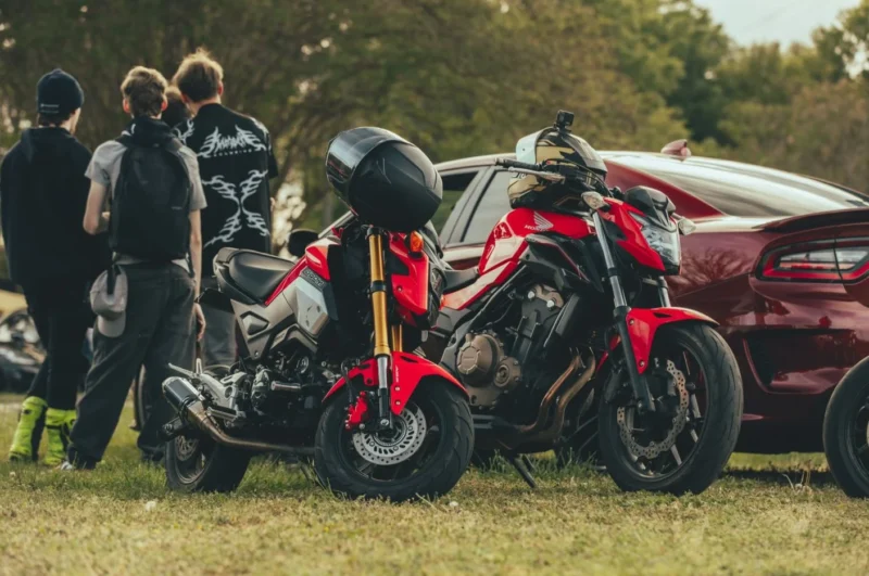 Red Honda motorcycle and red car at outdoor gathering with attendees nearby.