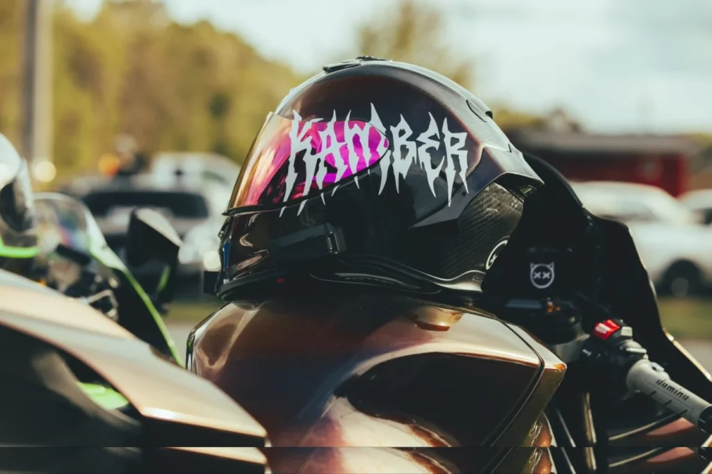 A close-up of a motorcycle helmet with a reflective visor, showing the word 'KAMBER' in a unique font, parked near other vehi
