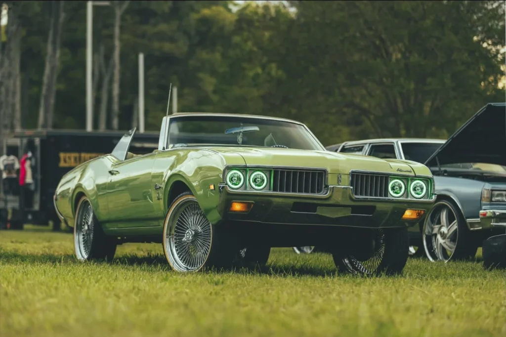 Green convertible muscle car with chrome accents parked on grass, other vehicles in background.