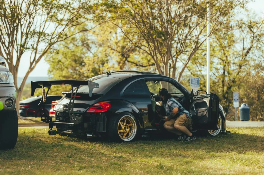 A black modified Volkswagen Beetle with an oversized rear wing parked on grass. A person leans into the car interior.
