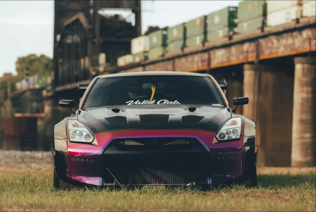 Nissan GT-R in a multicolored wrap parked on grass with a bridge in the background.