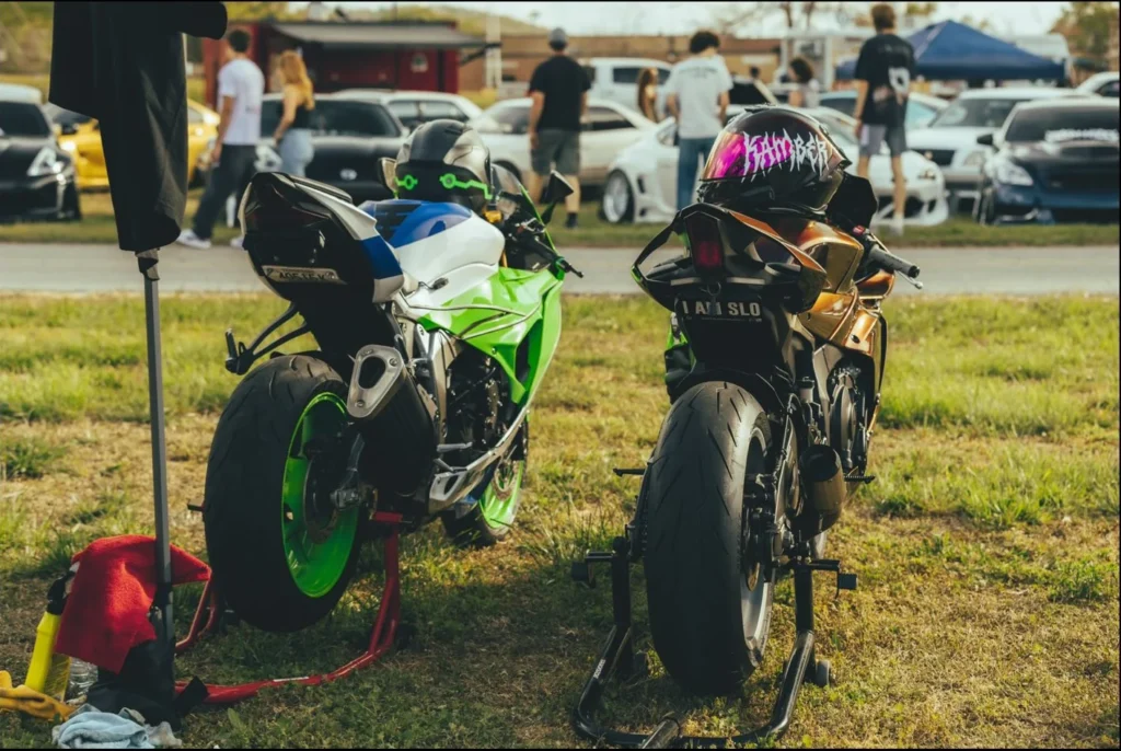 Two motorcycles, one green and white, the other gold, parked on grass with a crowd in the background.