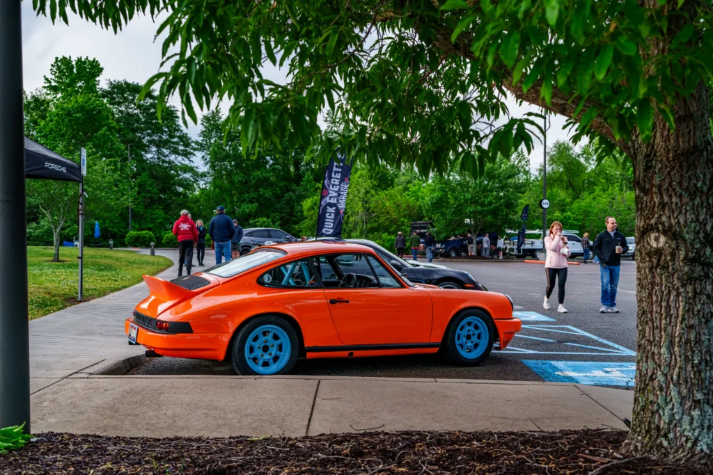 Orange Porsche 911 with blue wheels parked beside a sidewalk under tree branches at a car event