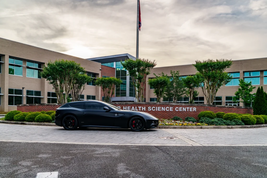 Black Ferrari FF parked in front of a brick sign and a multi-story building with trees around.