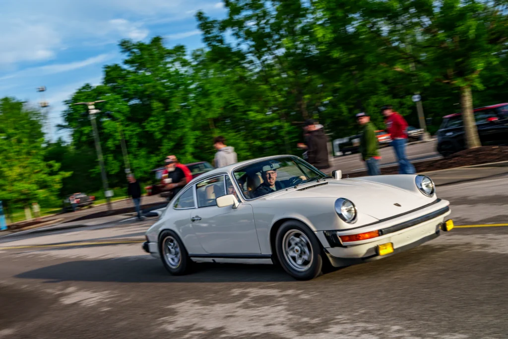 White Porsche 911 driving past a group of people in a parking lot with green trees and blue sky in the background.