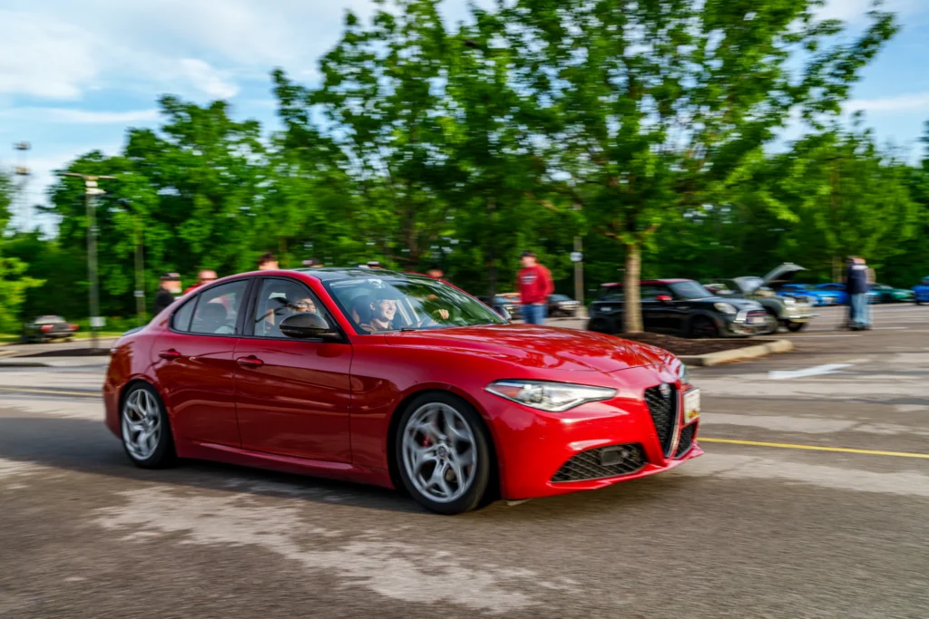 Red Alfa Romeo sedan driving on a street with people and parked cars in the background