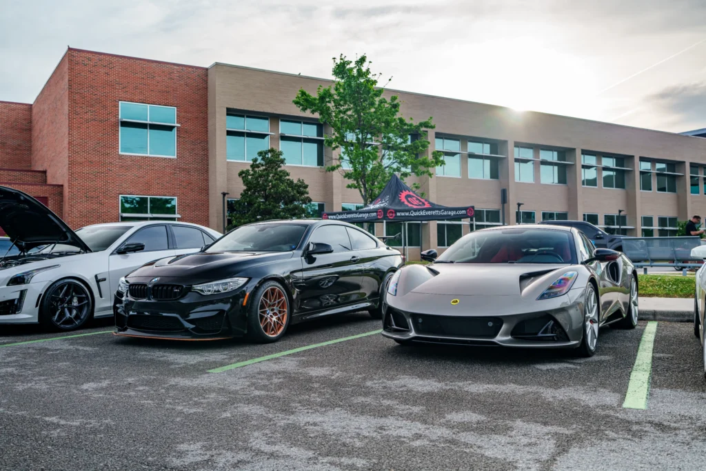 Black BMW M4 and silver Lotus Emira parked side by side in a lot at a car event with a building and tent in the background