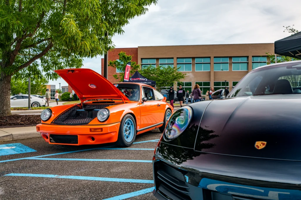 Orange Porsche 911 with front trunk open and black Porsche next to it at an outdoor car event in a parking lot