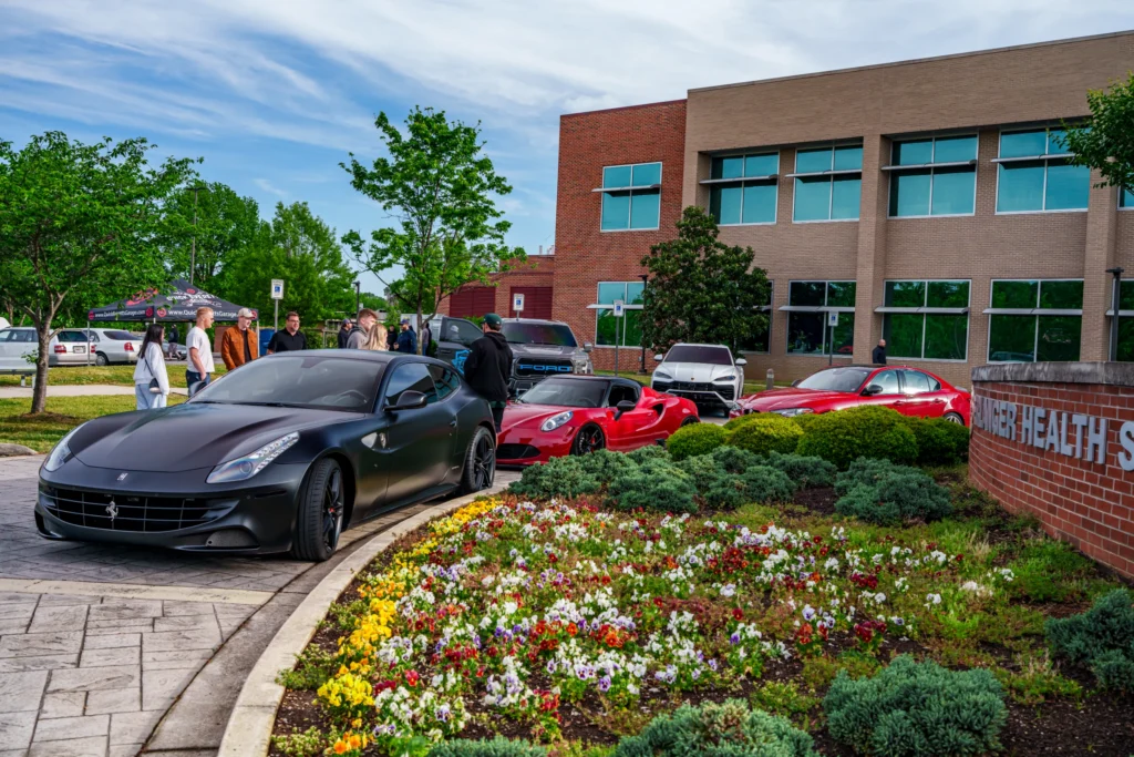Matte black Ferrari FF parked by a landscaped flower bed with people standing nearby and other vehicles in a parking lot