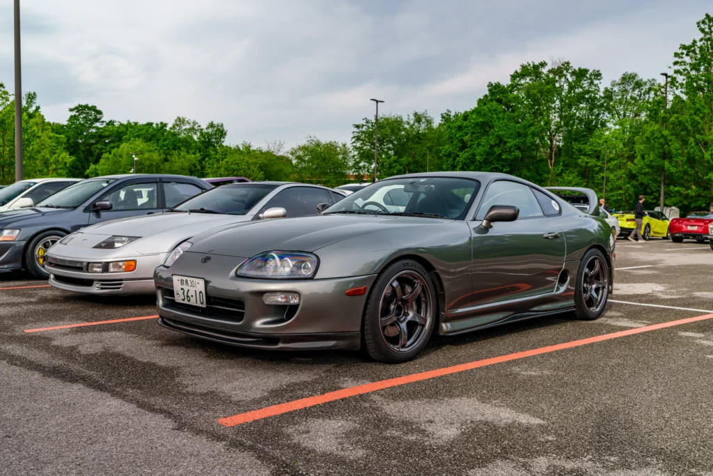 Silver Toyota Supra parked in a lot next to other cars with trees and cloudy sky in background