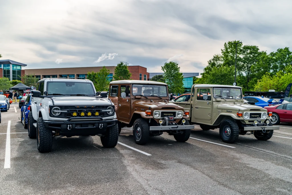 White Ford Bronco and two vintage Toyota Land Cruisers parked side by side at a car event in an outdoor lot