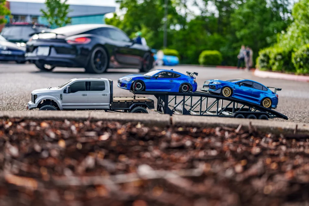 White RC car hauler with blue race car and blue sedan carrying ramps in a parking lot with other cars and trees in the backgr