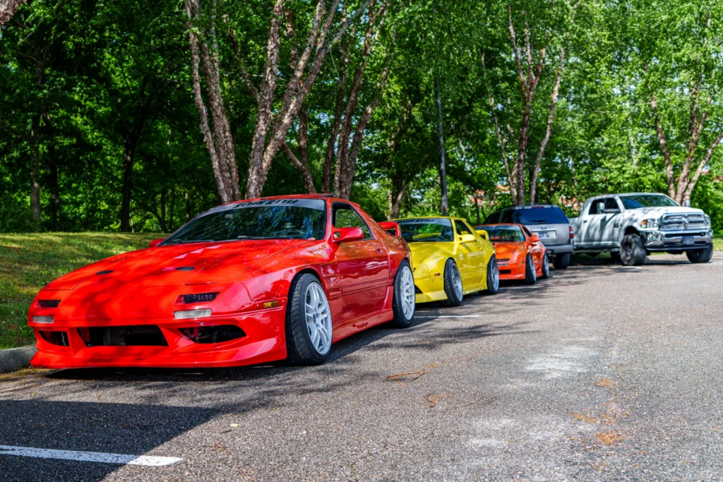 Red, yellow, and orange Mazda RX-7 cars parked in a line on a street with trees in the background