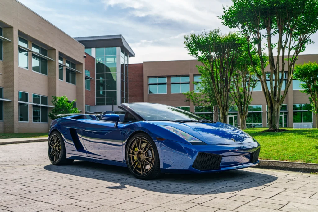 Blue Lamborghini Gallardo Spyder parked on paved area with modern building and trees in background