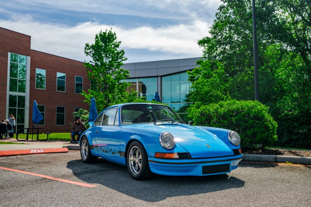 Blue Porsche 911 Carrera parked near a modern building with trees and people sitting in the background