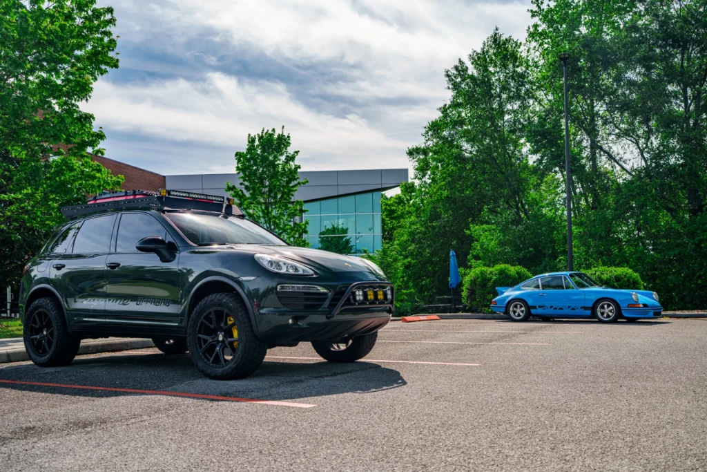 Black Porsche Cayenne Offroad and blue Porsche 911 Carrera parked in an outdoor lot with trees and modern building in backgro
