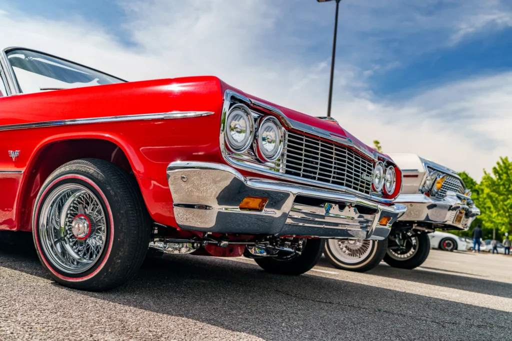Red Chevrolet Impala with wire-spoke wheels parked next to other classic cars under a partly cloudy sky