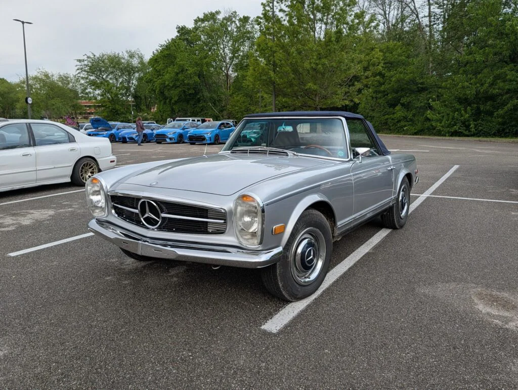 A silver convertible Mercedes-Benz parked in a lot with other cars and trees in the background.