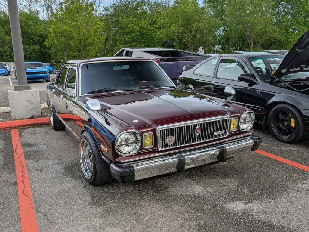 A maroon Toyota Cressida with chrome accents parked on a lot, surrounded by other vehicles and greenery in the background.