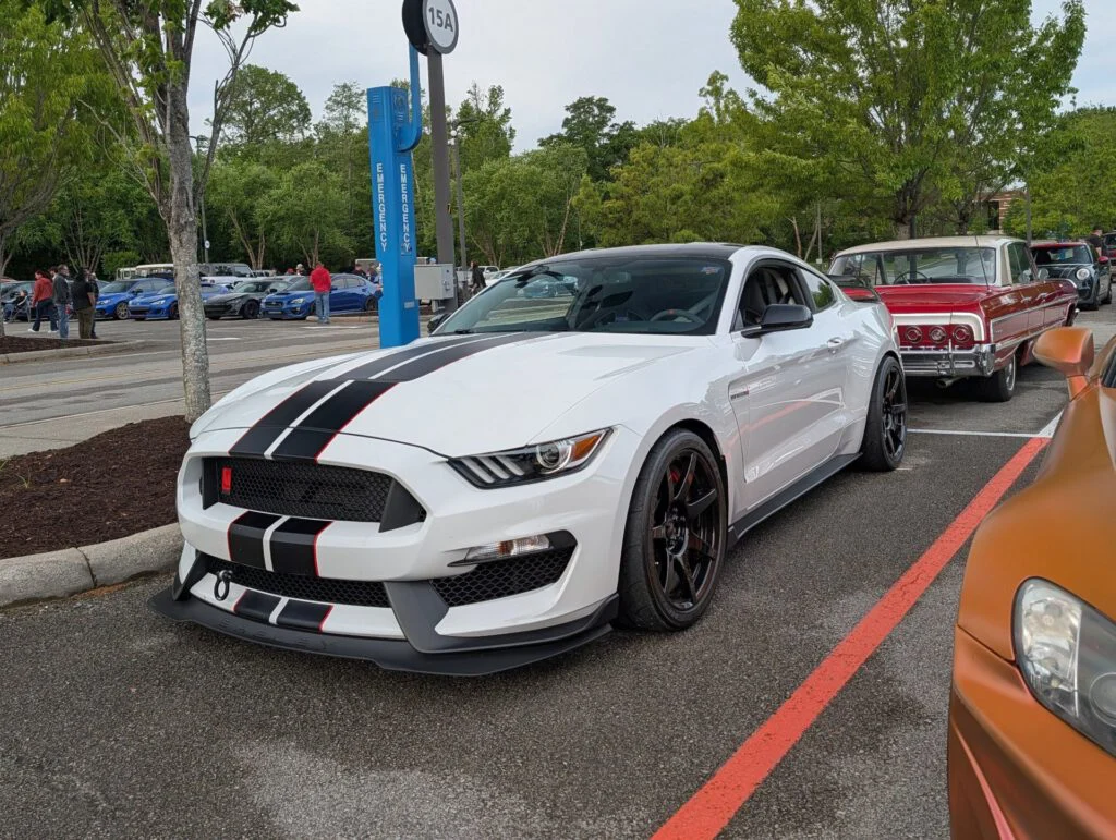 A white Ford Mustang with black stripes parked beside a vintage red Chevrolet in a gathering area with trees and other cars i