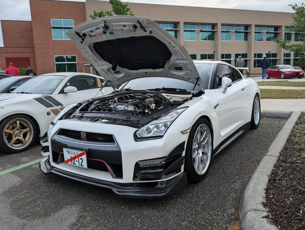 White Nissan GT-R with the hood raised, revealing the engine, parked next to another vehicle at a car event.
