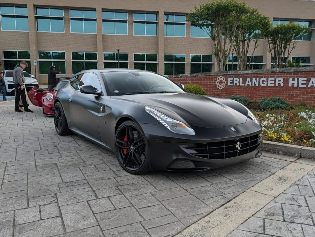 Matte black Ferrari FF parked outside a building with a brick wall and flower beds. People are visible in the background.
