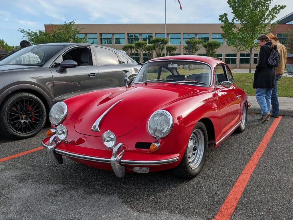 Red Porsche 356 parked beside a gray SUV in a lot with trees and a building in the background.