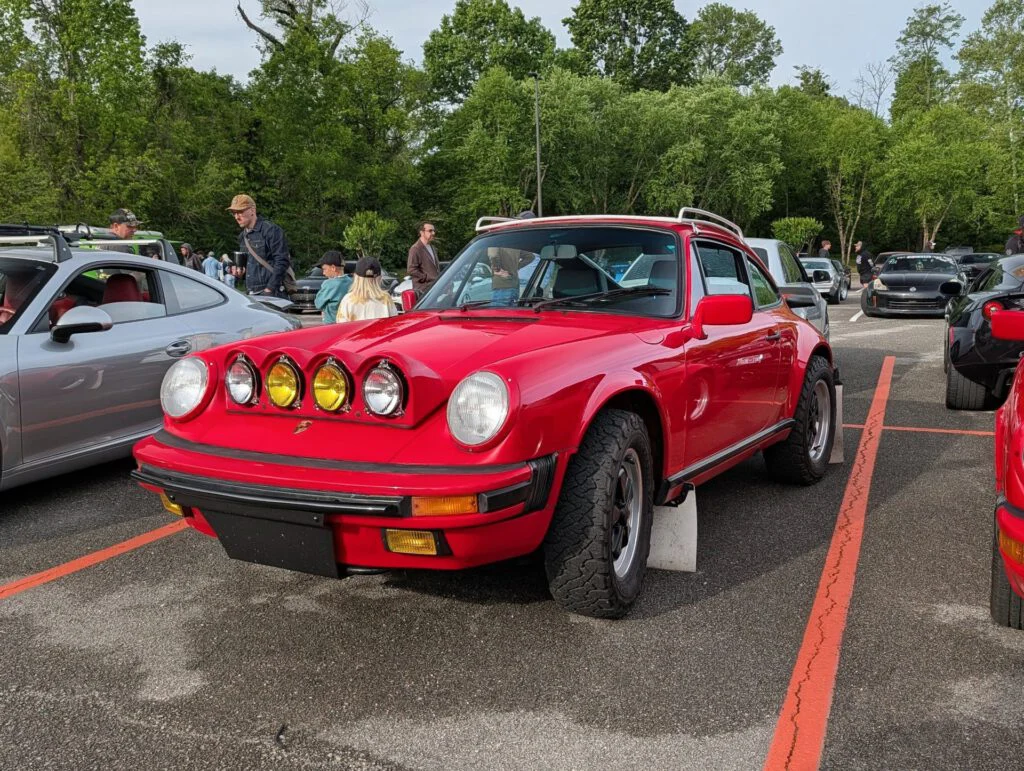 Red Porsche 911 with rally lights parked at a car event, surrounded by attendees and other vehicles in a lot.