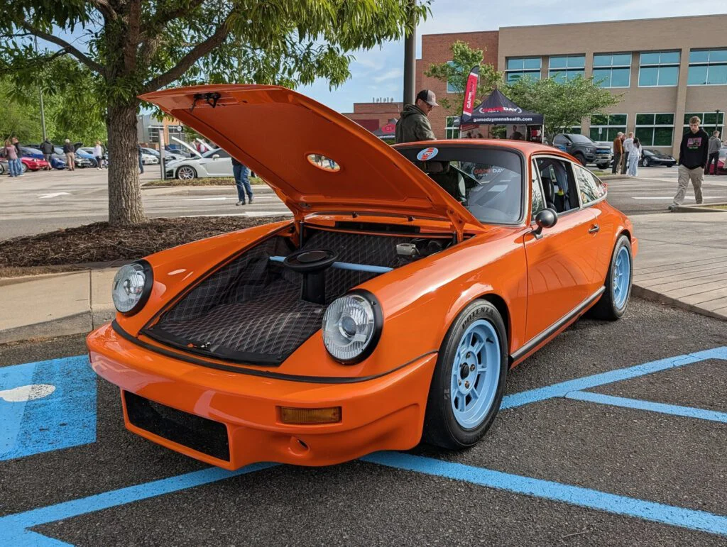 Orange sports car with hood raised, displaying engine bay. Attendees and other vehicles in the background at a car event.